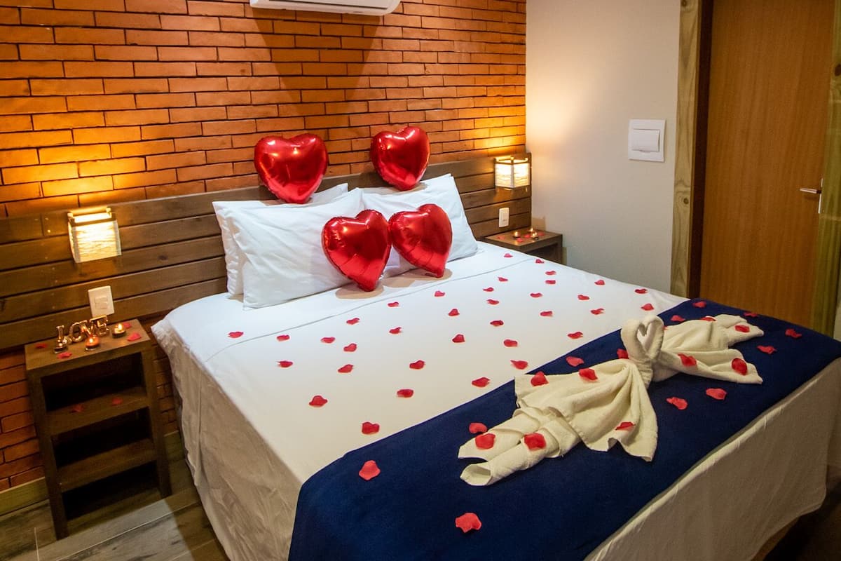 A hotel room with a brick wall, wooden beams, a bed with white bedding, red heart-shaped balloons, and a blue blanket with rose petals.