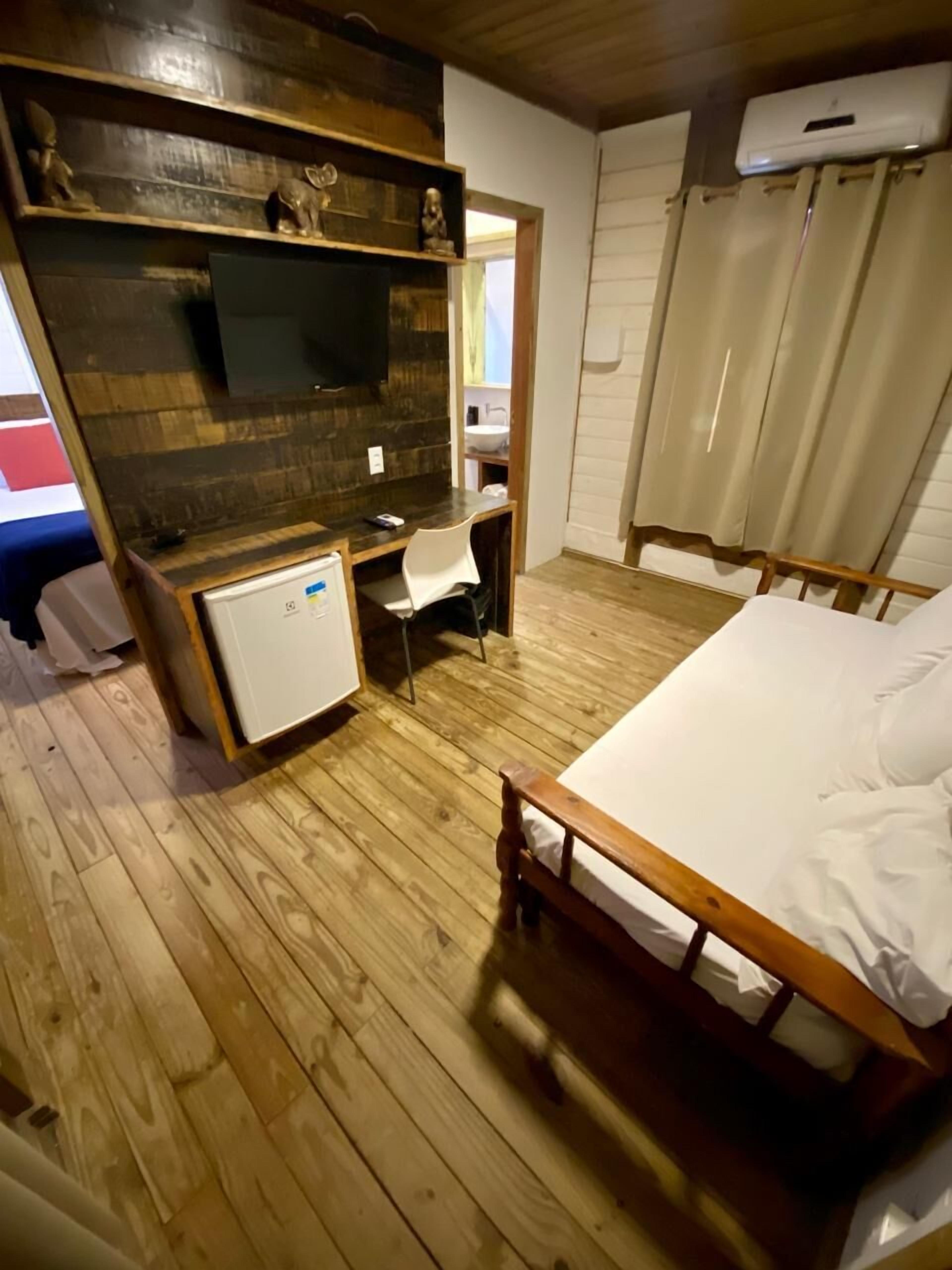 A bedroom with a wooden floor, a bed with white linens, a desk with a chair, a small white cabinet, a television on the wall, and a window with curtains.
