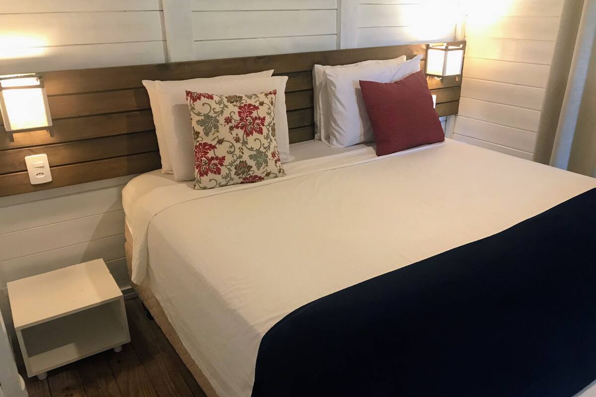 A neatly made bed with a white duvet, a floral pillow, and a red pillow. There is a small bedside table with a lamp on it.