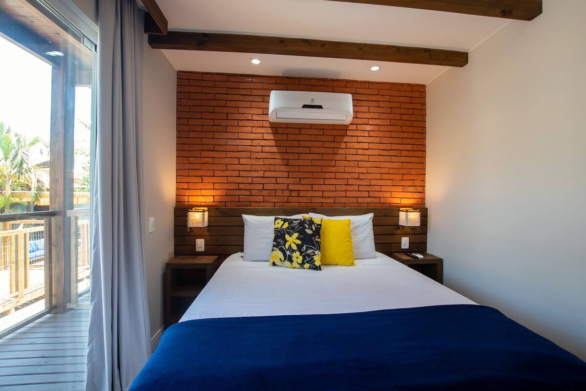 A hotel room with a brick wall, a bed with white and blue bedding, two bedside lamps, and an air conditioning unit.