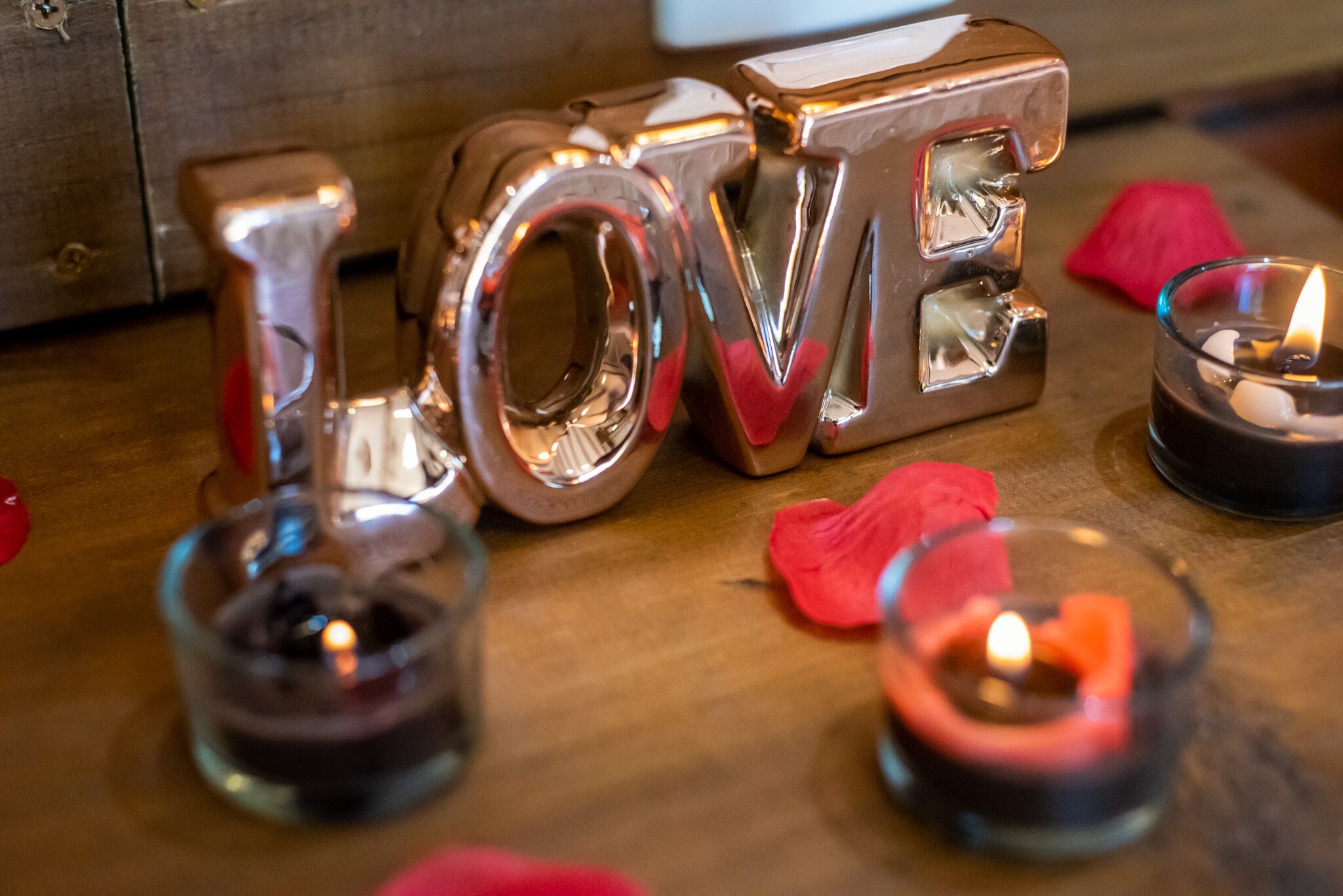 Letras metálicas formando a palavra 'LOVE' sobre uma superfície de madeira, com velas e pétalas de rosa.