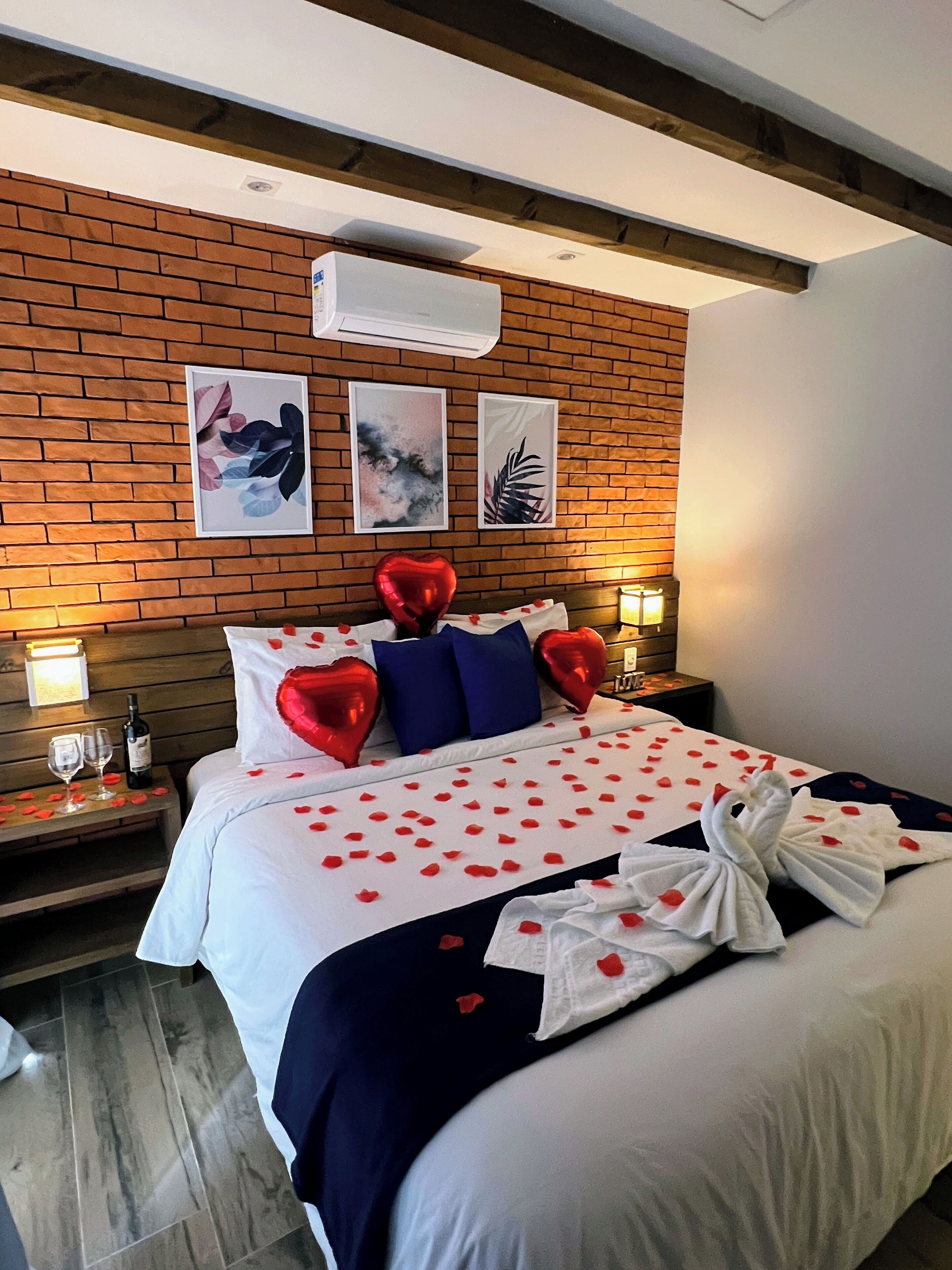 Una habitación de hotel con una pared de ladrillo, una cama decorada con globos rojos con forma de corazón, un ramo de rosas rojas y una botella de vino en la mesita de noche.