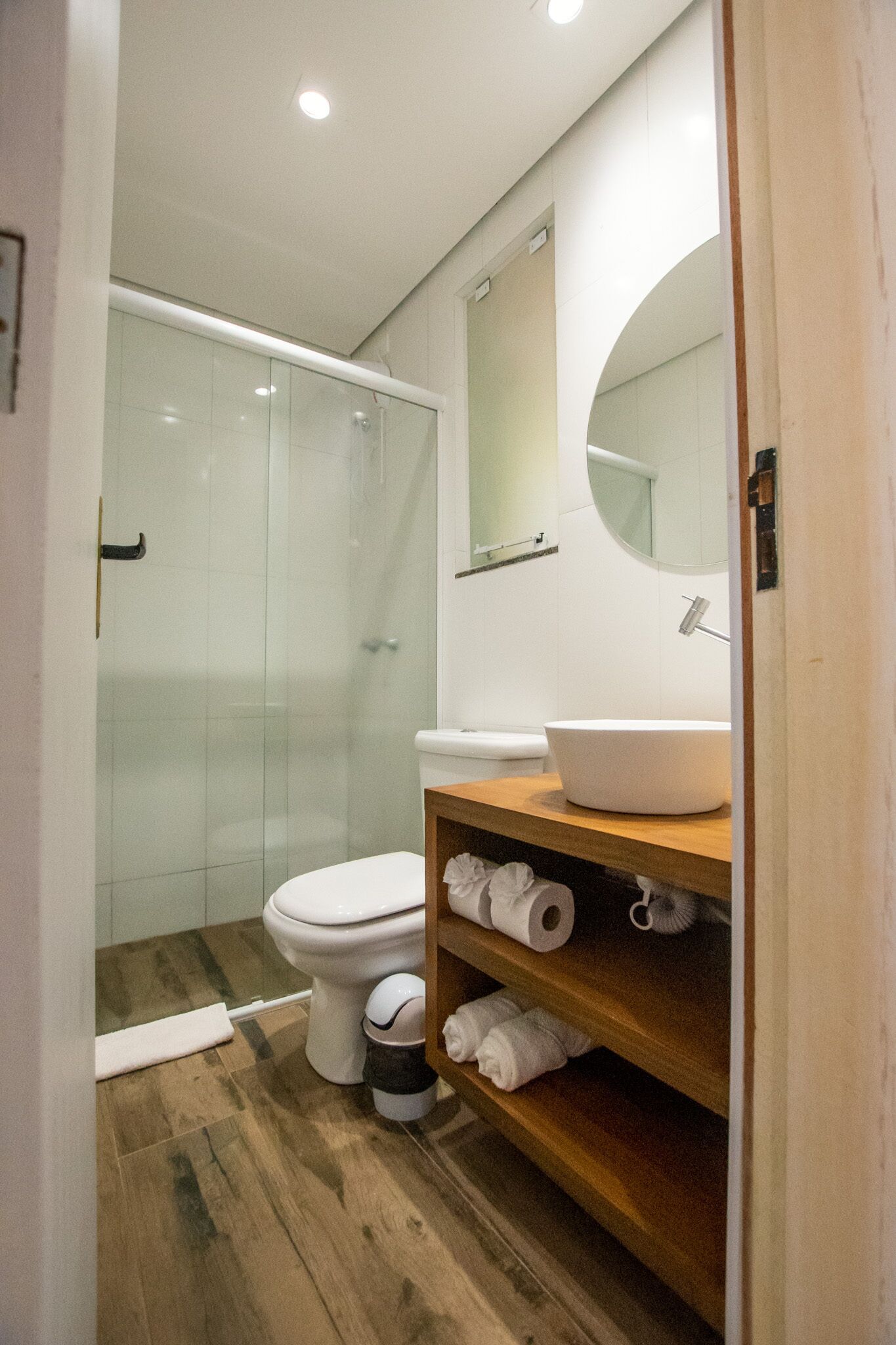 Un baño moderno con cabina de ducha de vidrio, inodoro blanco, tocador de madera con lavamanos grande y espejo sobre el lavamanos.