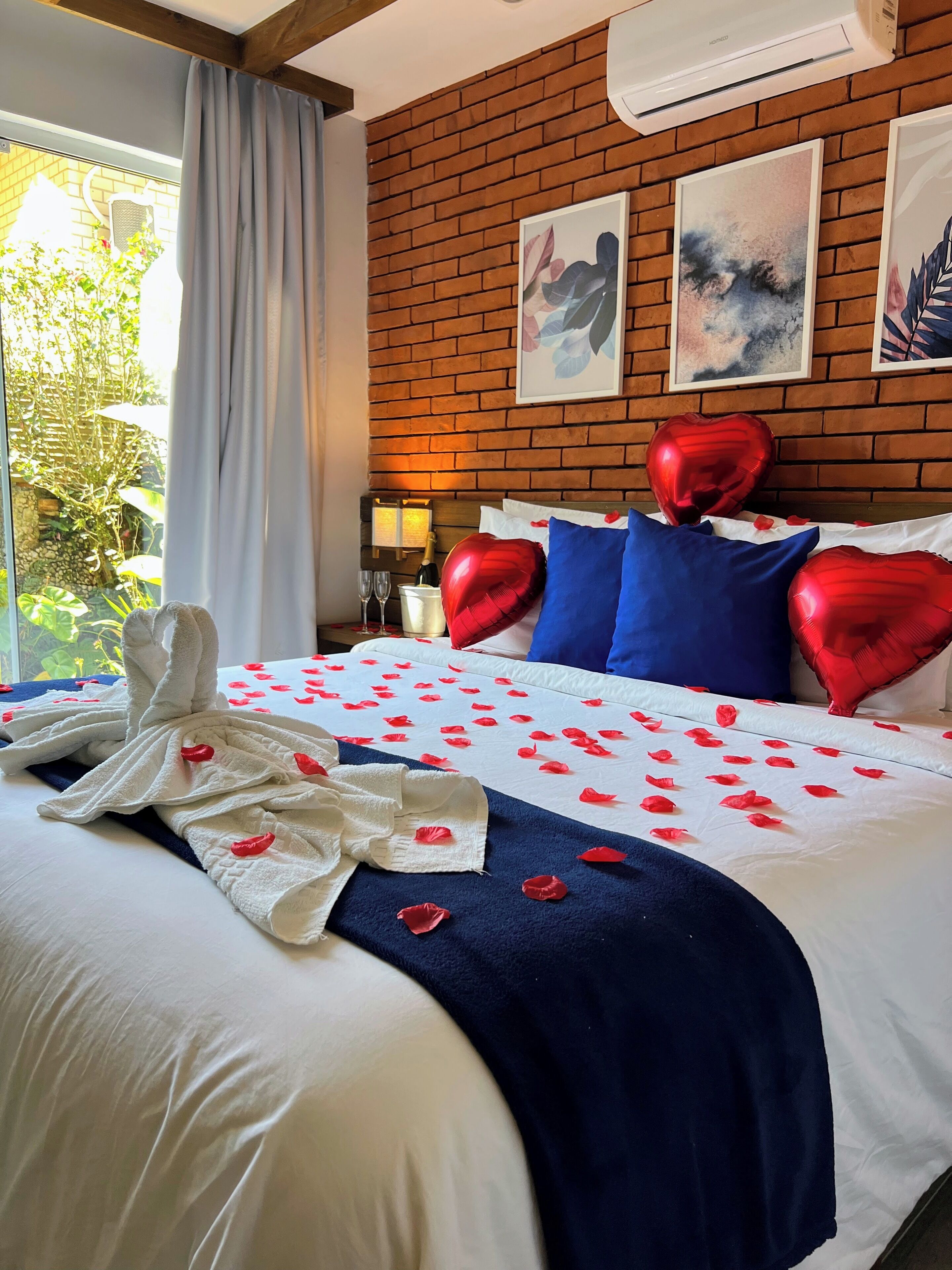A hotel room with a brick wall, a bed with red heart-shaped pillows, a white bedspread with red rose petals, and a white towel on the bed.