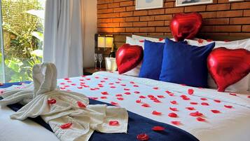 A hotel room with a brick wall, a bed with red heart-shaped pillows, a white bedspread with red rose petals, and a white towel on the bed.