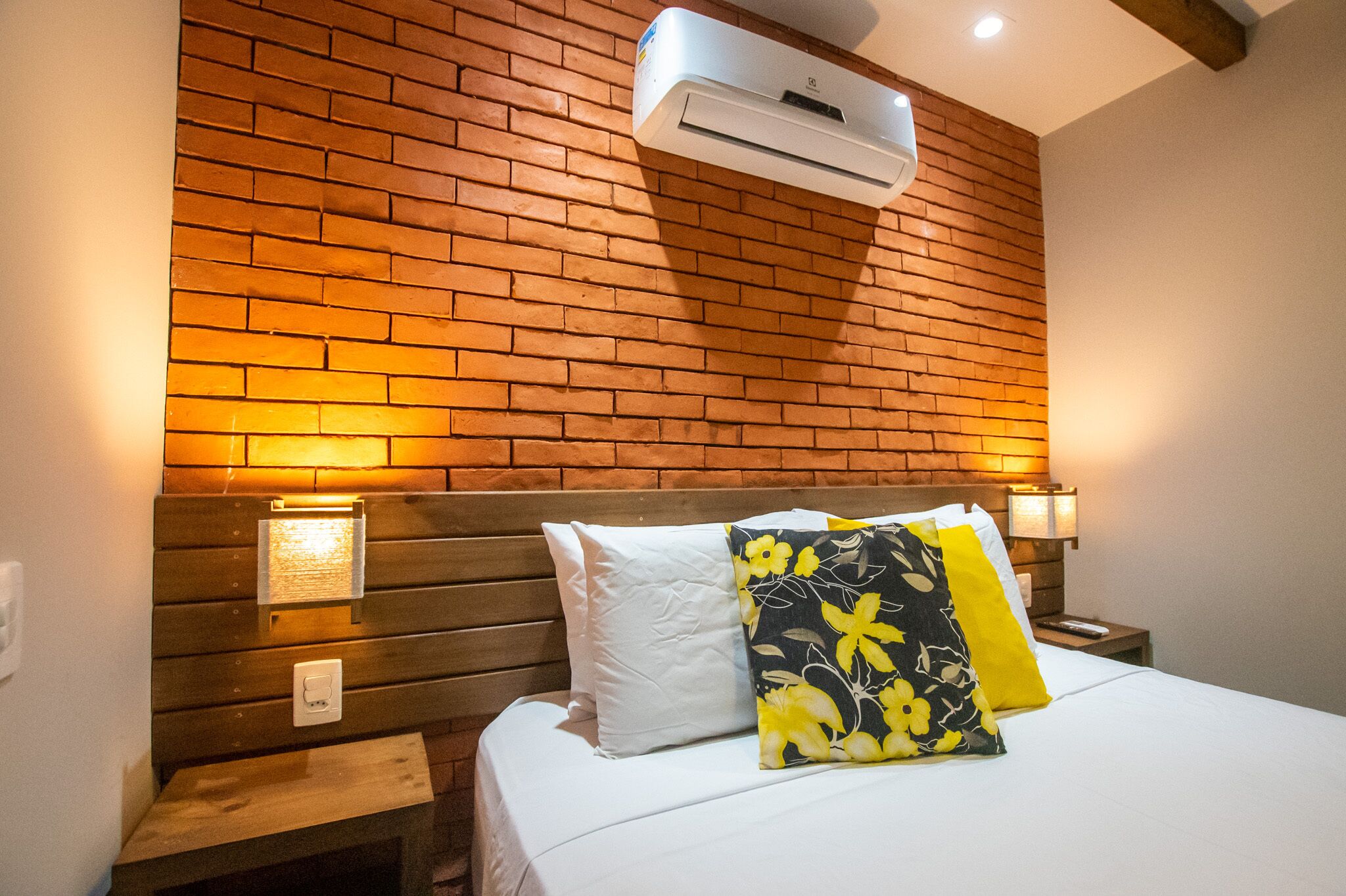Habitación de hotel con pared de ladrillo, cabecera de madera, cama con ropa blanca y almohada estampada con motivos florales.