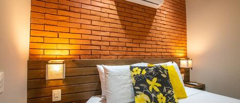 A hotel room with a brick wall, a wooden headboard, a bed with white bedding, and a floral pillow.