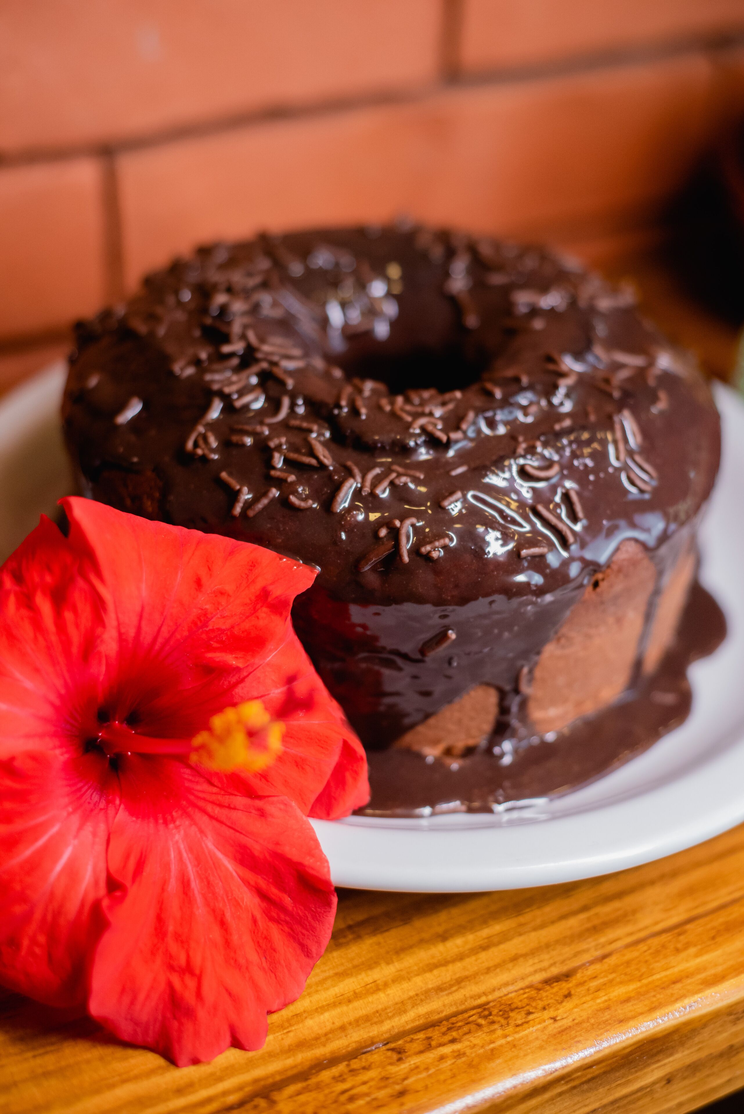 A chocolate-glazed donut with sprinkles on a white plate, accompanied by a red hibiscus flower.