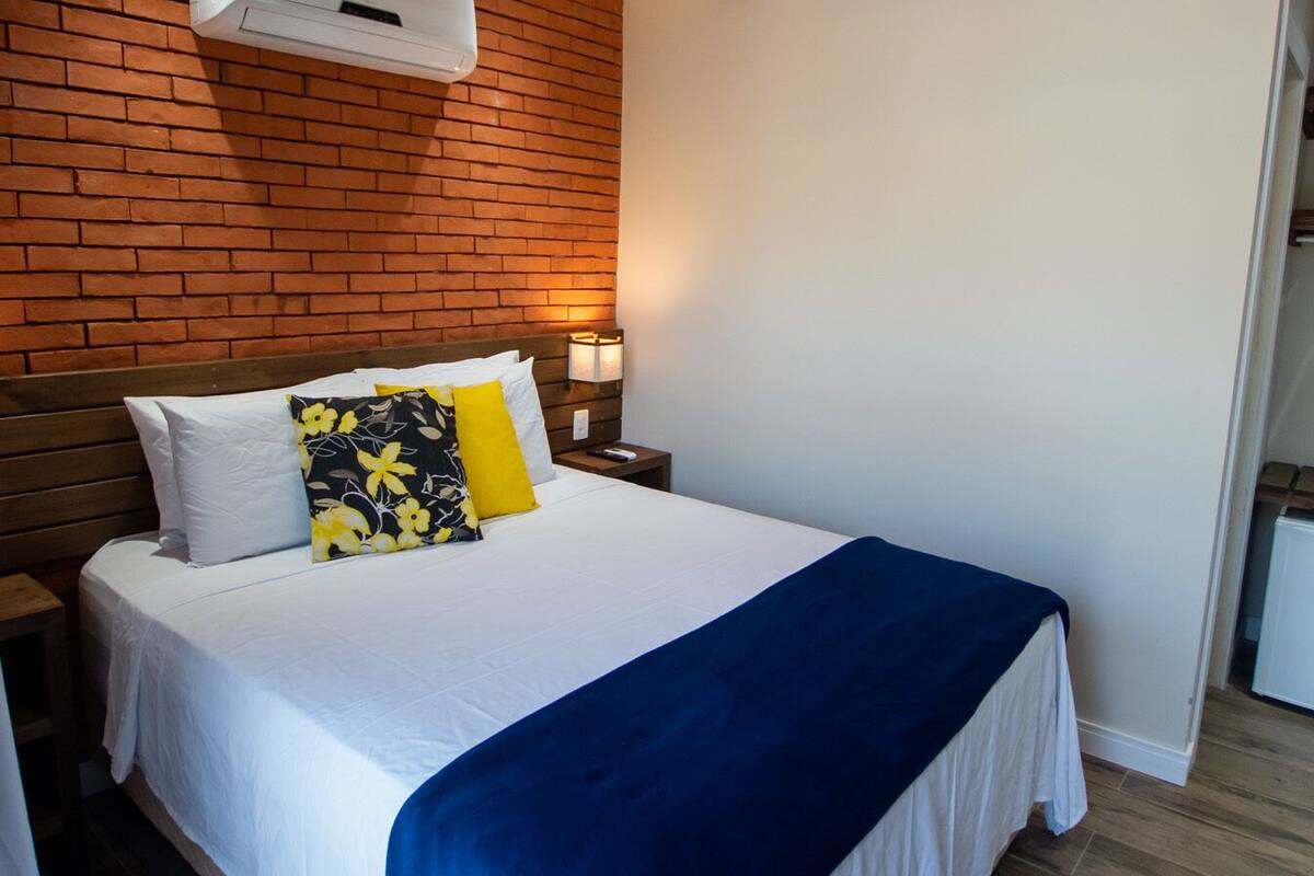 A hotel room with a brick wall, a bed with white bedding and a blue blanket, a nightstand with a lamp, and a ceiling fan.