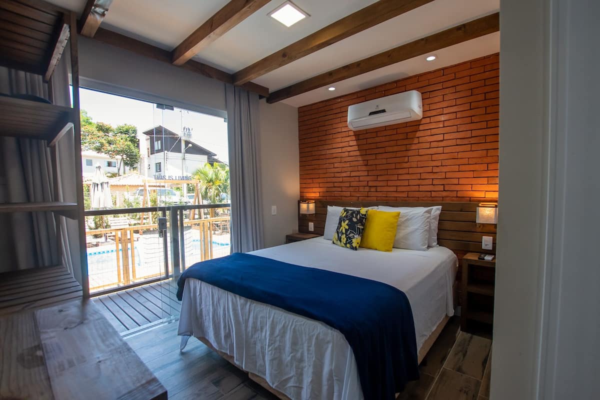 A bedroom with a large bed, a wooden ceiling, a brick wall, and a balcony with a view of the outside.