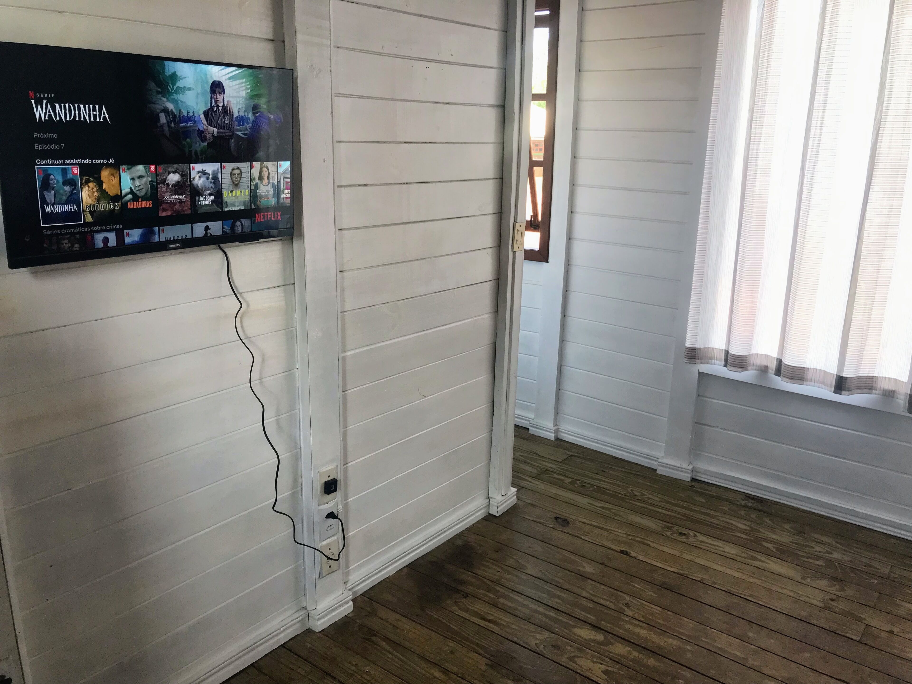 A room with white paneled walls, a wooden floor, and a flat-screen TV mounted on the wall displaying a Netflix series.