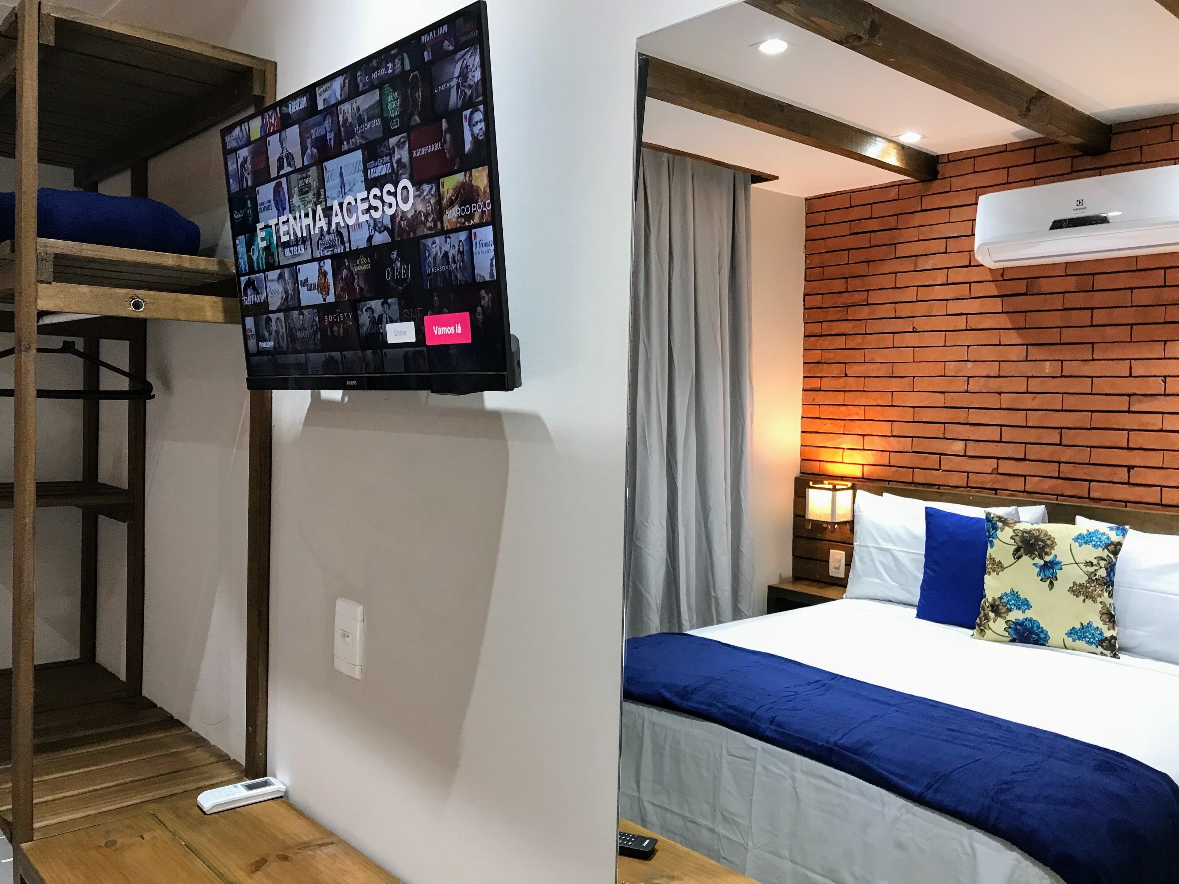 A hotel room with a TV displaying movie posters, a bunk bed, a brick wall, and a bed with blue and white bedding.