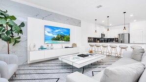 A modern living room with a white sofa, a glass coffee table, and a large TV mounted on the wall. - EV_3009 - 9BR / 5BA Storey Lake Villa near to Disney, Private Pool & Games Room (Kissimmee)