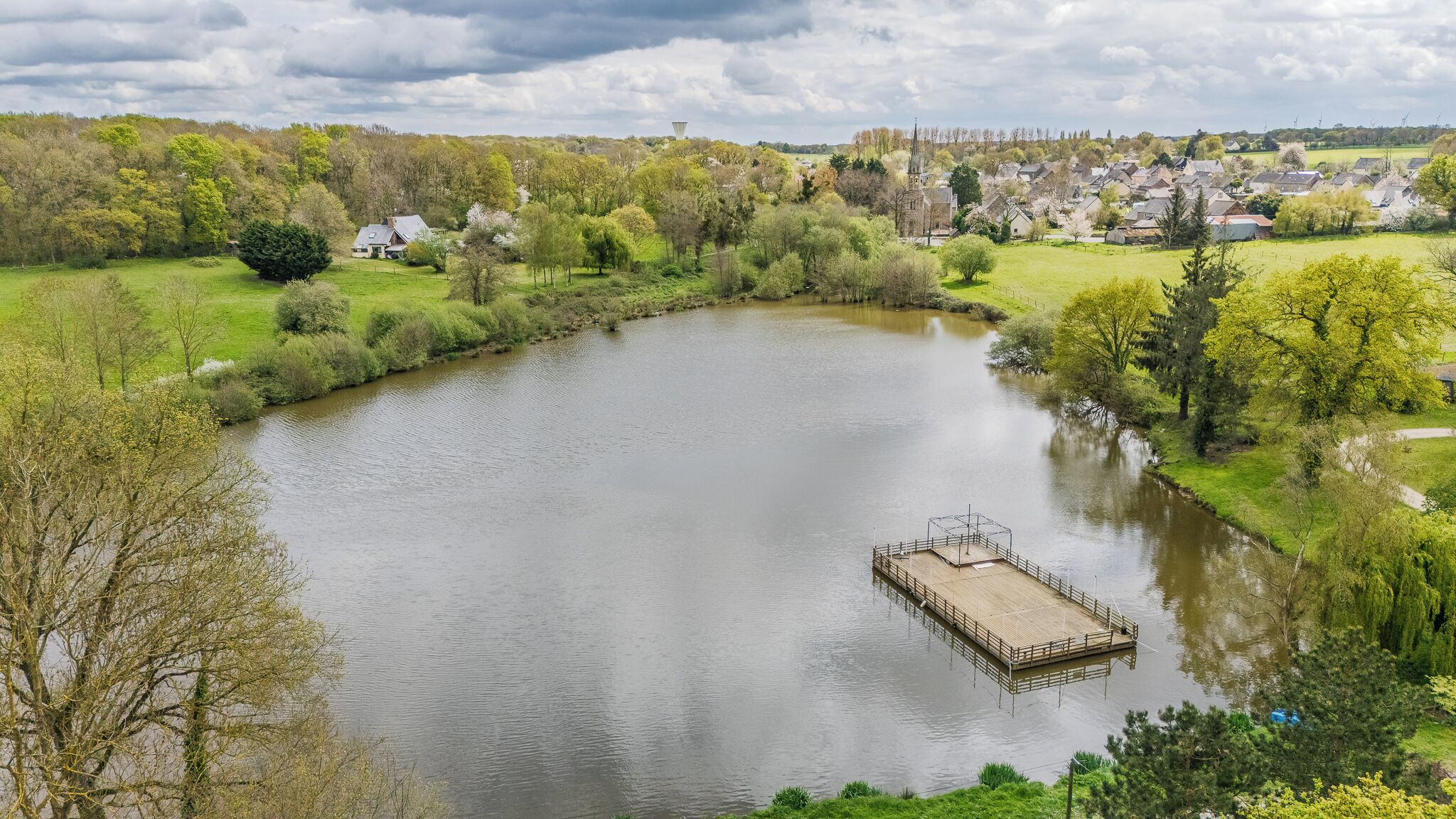 A serene lake with a dock, surrounded by lush greenery and a village in the background.