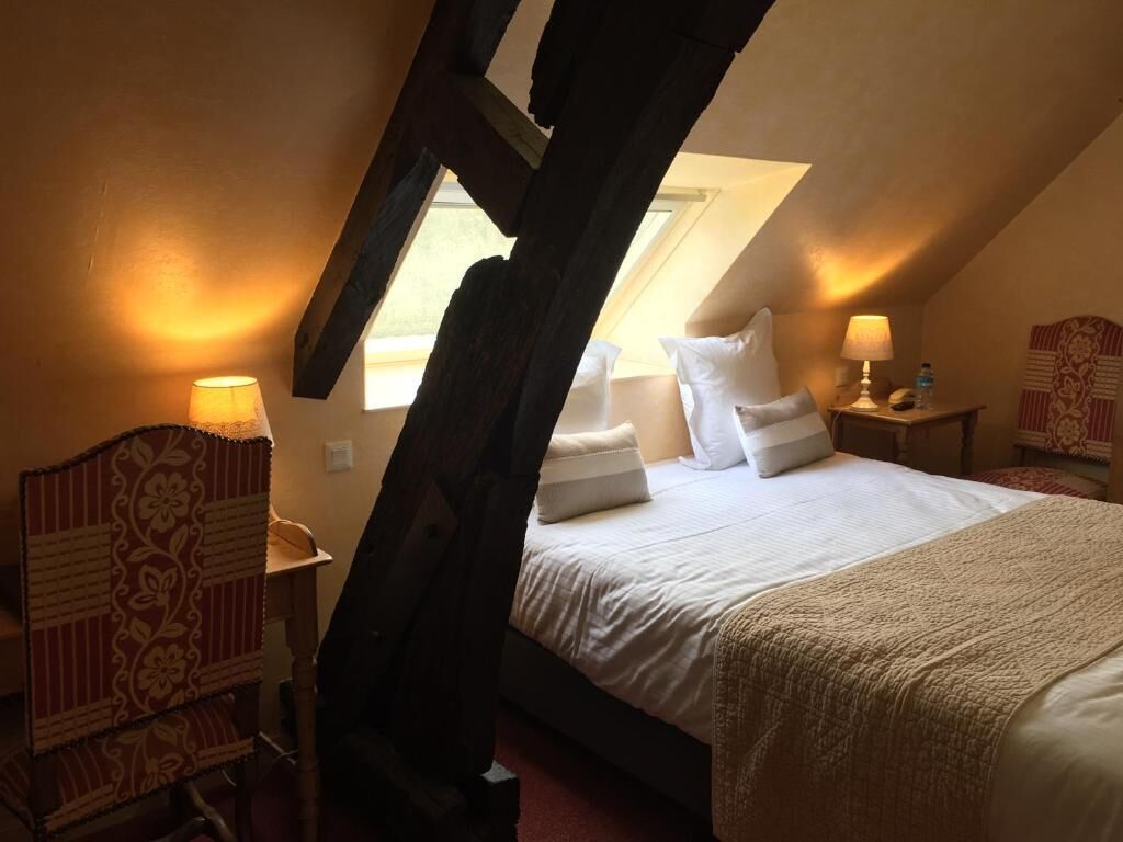 A bedroom with a sloped ceiling, a bed with white bedding, a wooden nightstand, a lamp, and a patterned chair.