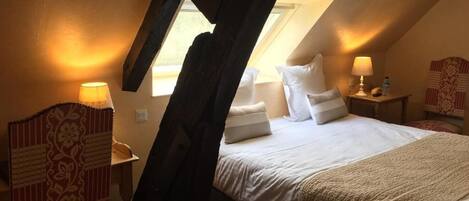 A bedroom with a sloped ceiling, a bed with white bedding, a wooden nightstand, a lamp, and a patterned chair.