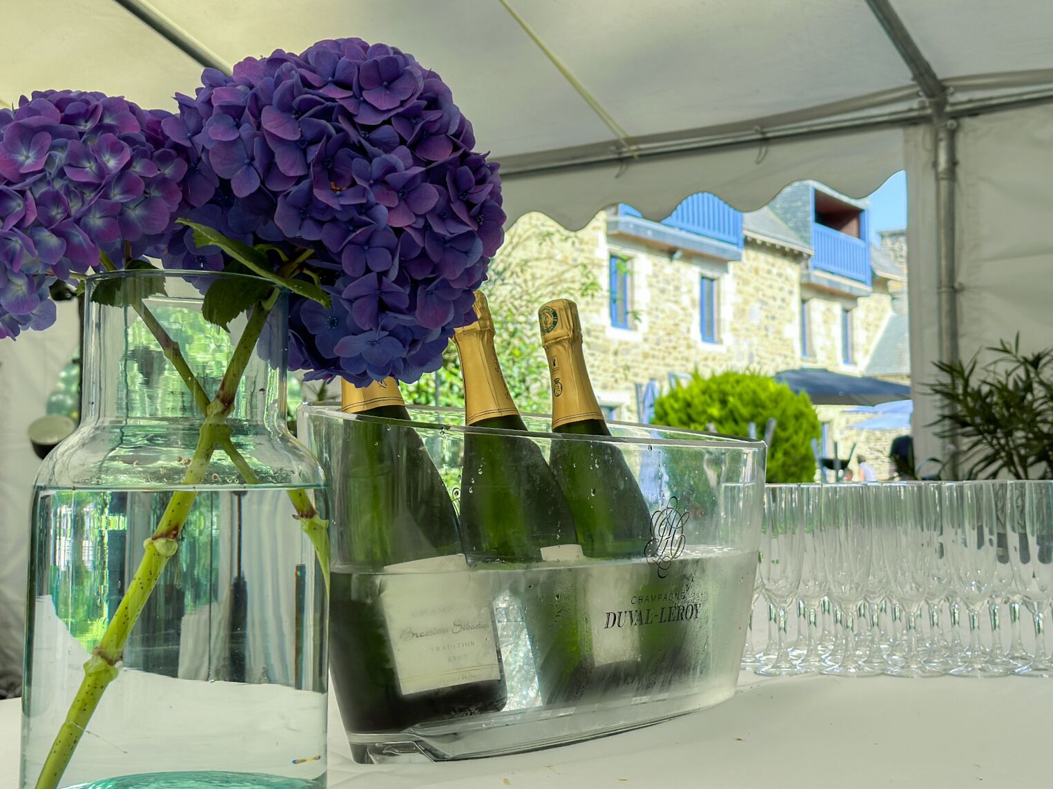 Een glazen vaas met champagneflessen en paarse bloemen.