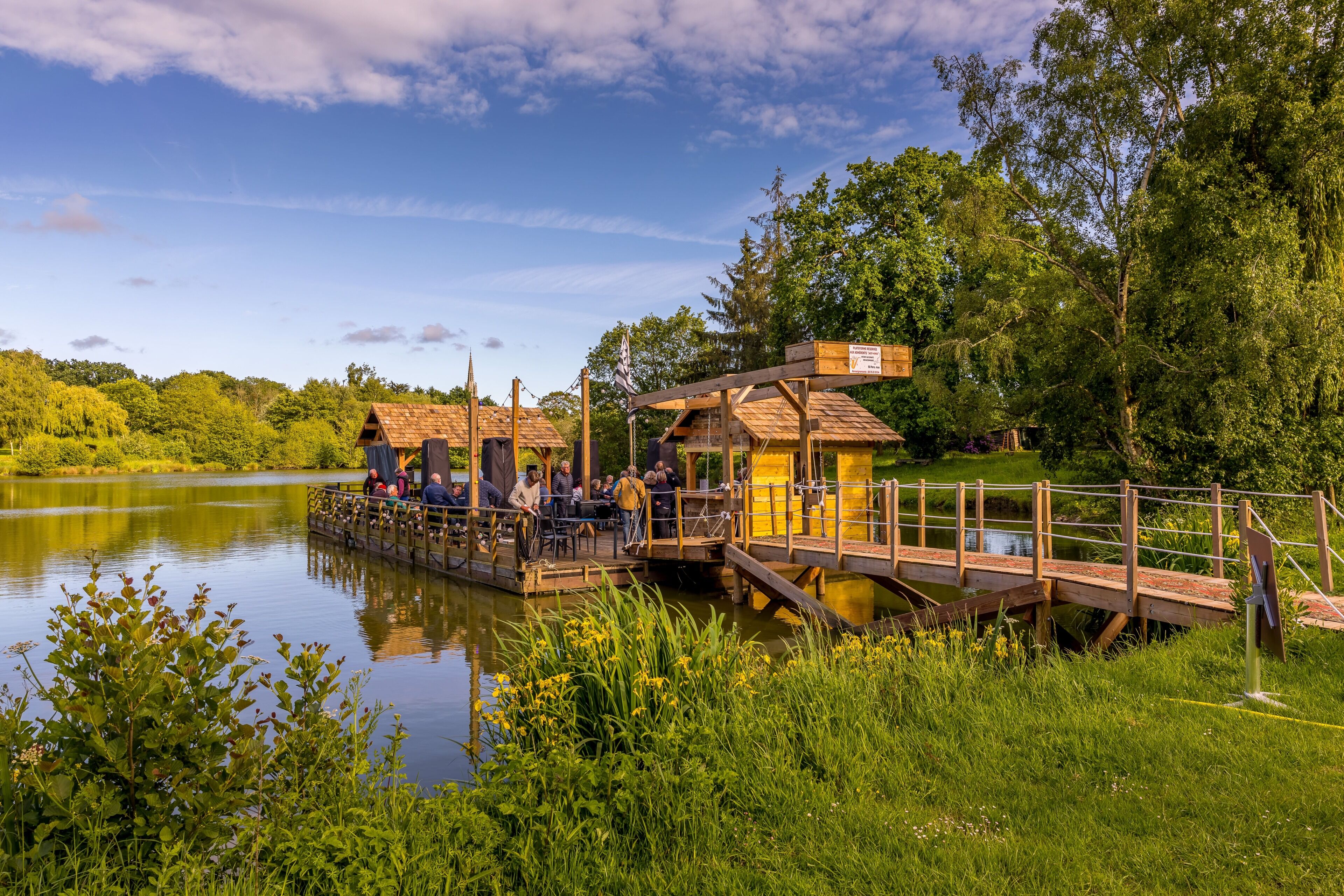 A wooden dock with a small building, a covered seating area, and a bridge over a calm lake.