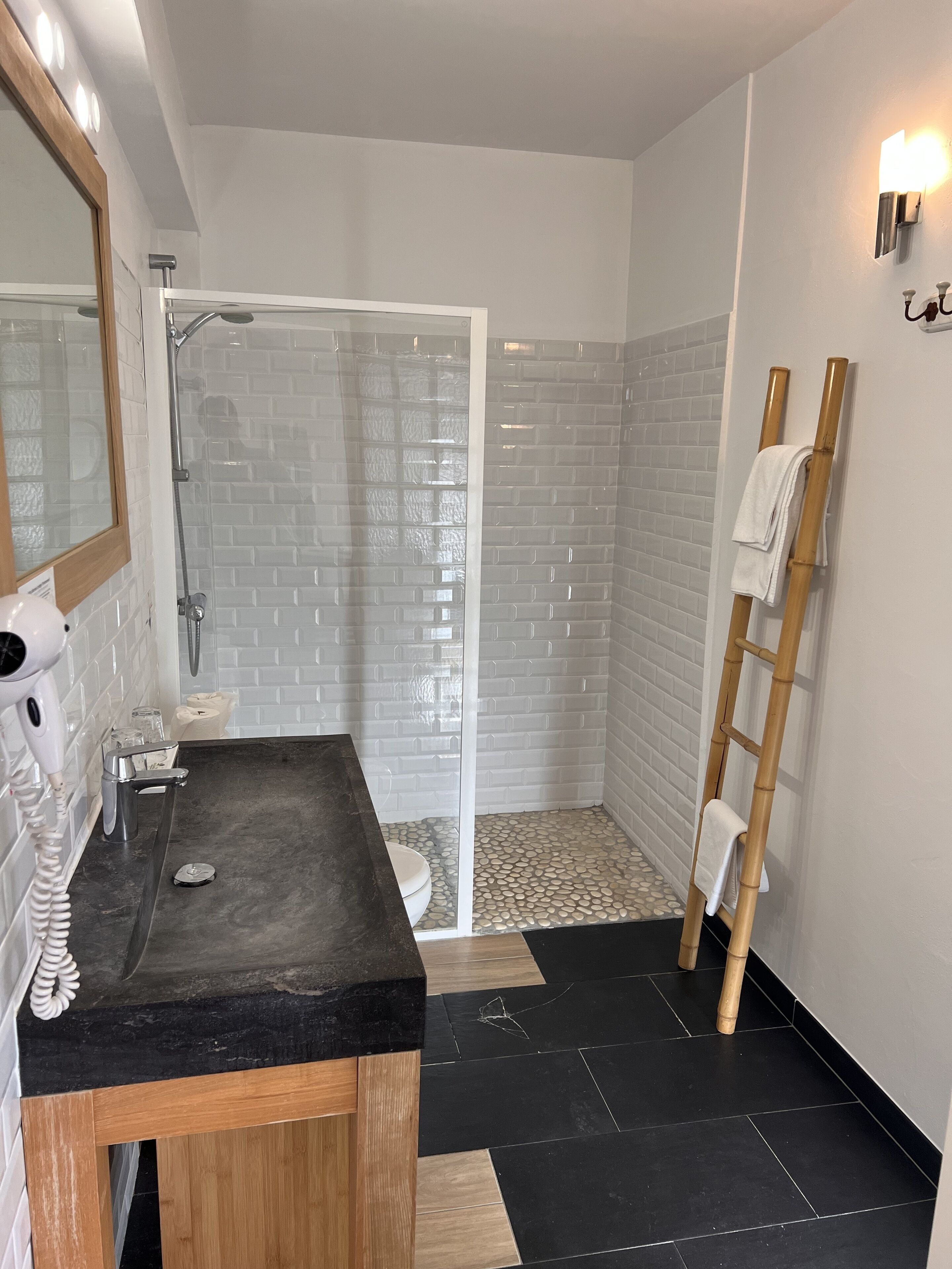 Un baño moderno con ducha envuelta en vidrio, tocador con encimera negra y un taburete de madera.
