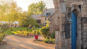 Un arc en pierre mène à un bâtiment doté d’une porte bleue, qui est entouré d’un jardin agrémenté d’un pot rouge et d’un banc.