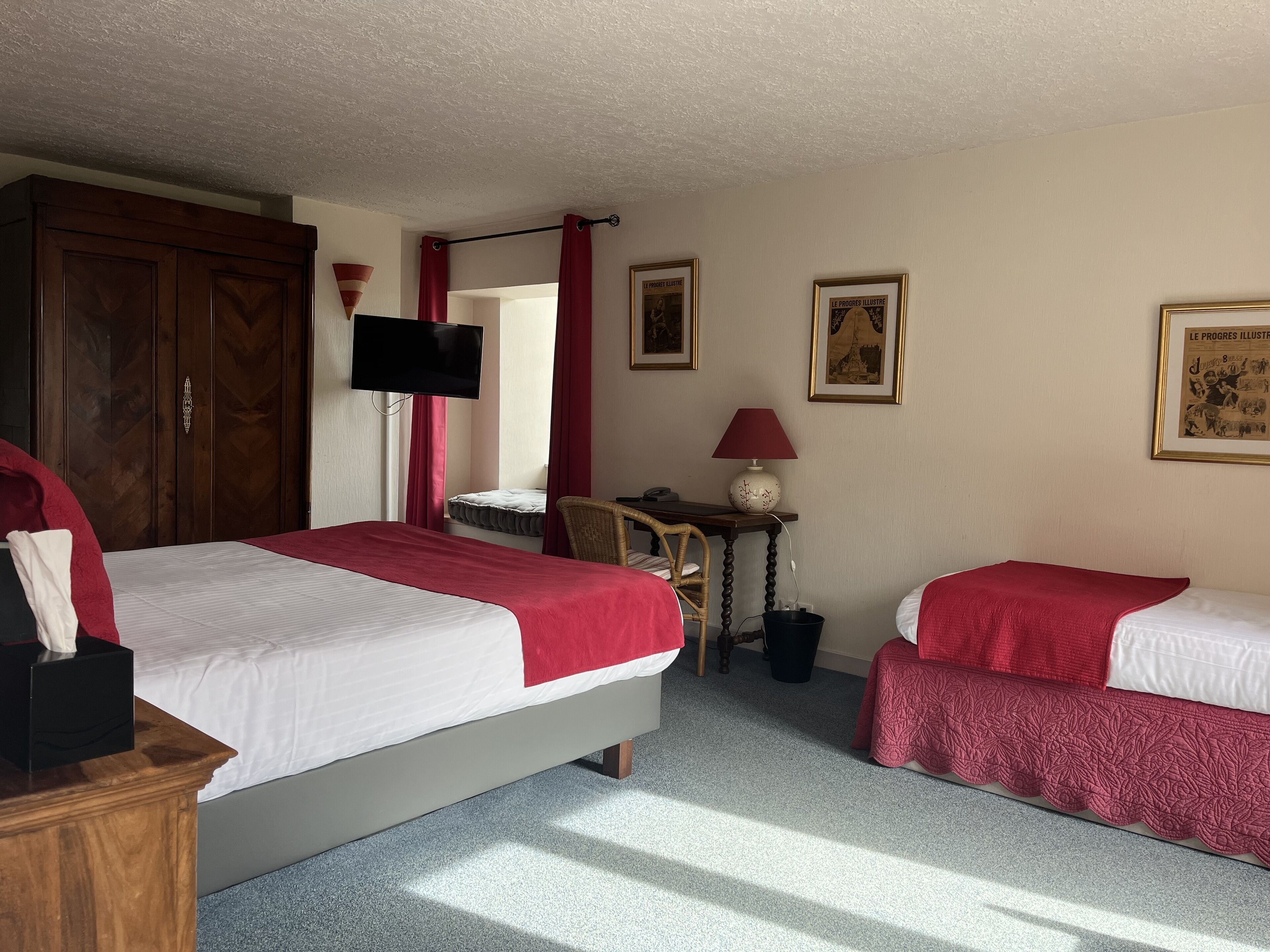 A hotel room with two beds, a wooden wardrobe, a television, a small table with a lamp, and framed pictures on the wall.