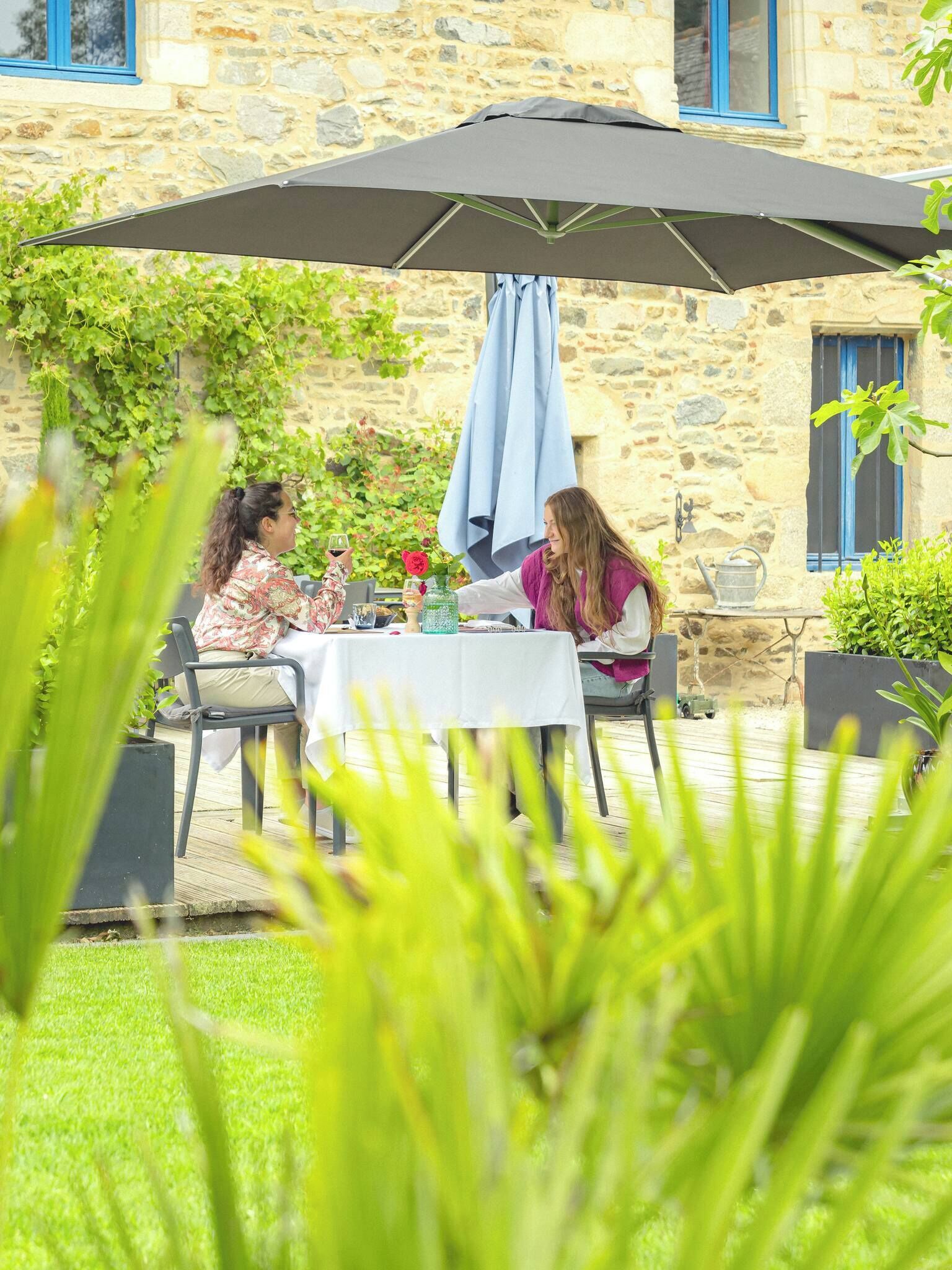 An outdoor dining area with a table and chairs under a large umbrella, set against a stone building with a garden.