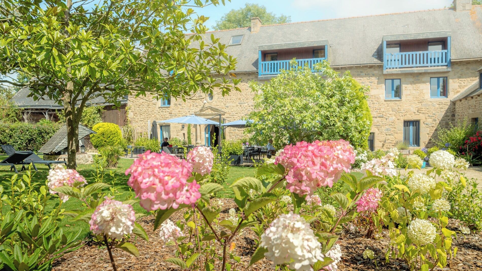 Un edificio de piedra con balcones azules, rodeado por un jardín lleno de flores rosas y blancas.