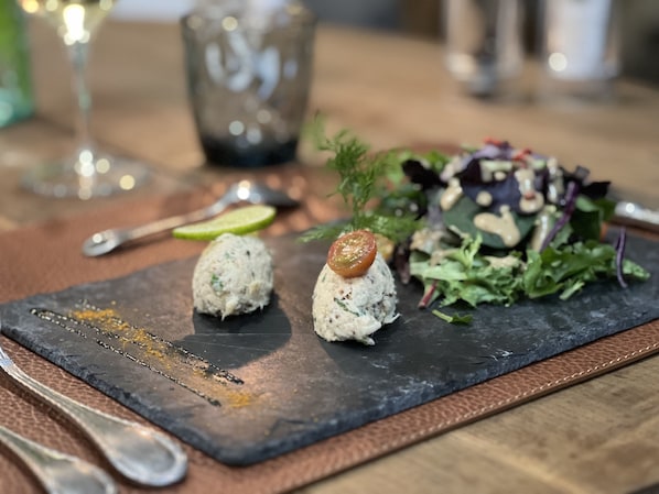 Une planche de service en ardoise sur laquelle reposent deux aliments de forme sphérique, une salade et un verre de vin blanc, le tout posé sur une table.