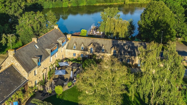 Vue aérienne d’un bâtiment historique en pierre, doté d’une cour et d’un espace extérieur aménagé pour s’asseoir, le tout entouré d’une végétation luxuriante et bordé d’un lac.