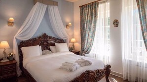 Un lit à baldaquin avec des draps blancs et un dais, deux tables de chevet avec des lampes, des rideaux à motifs floraux et un plancher en bois.