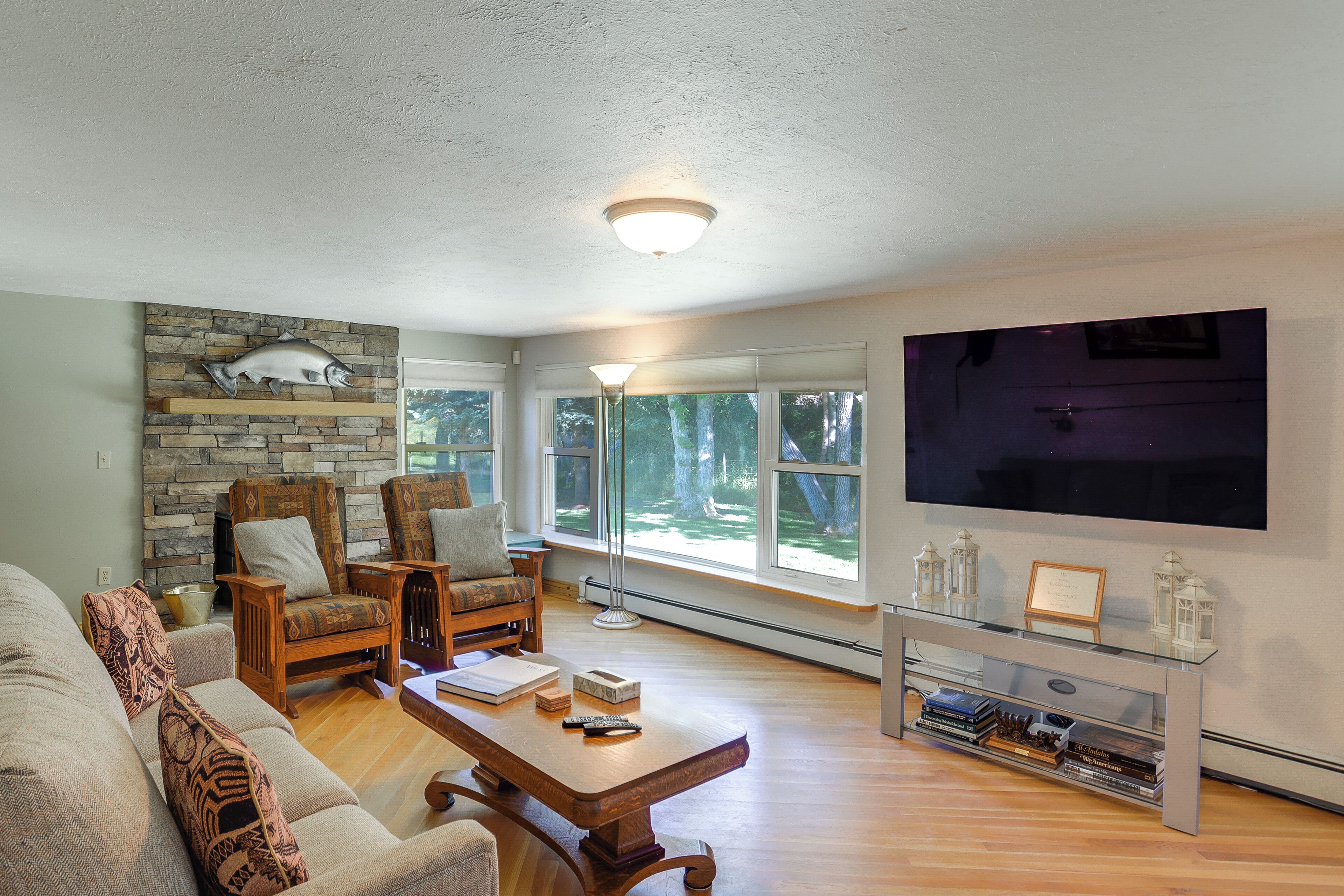 A living room with a stone fireplace, two patterned armchairs, a wooden coffee table, a flat-screen TV, and a bookshelf.