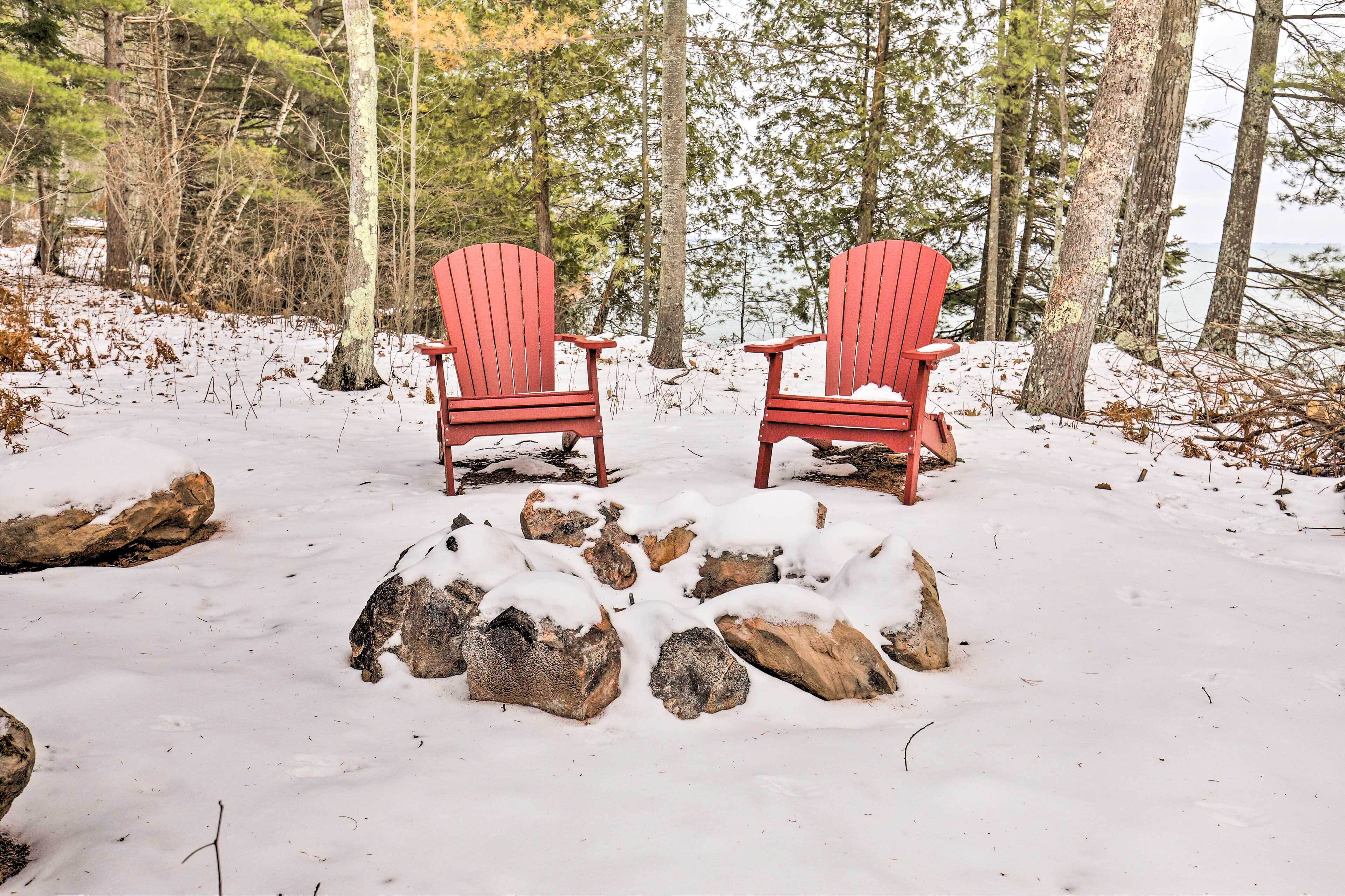 Two red Adirondack chairs facing a snow-covered log fire in a forested area.