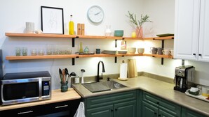 A kitchen with a microwave, sink, and coffee maker. There are shelves with various kitchen items and a clock on the wall.