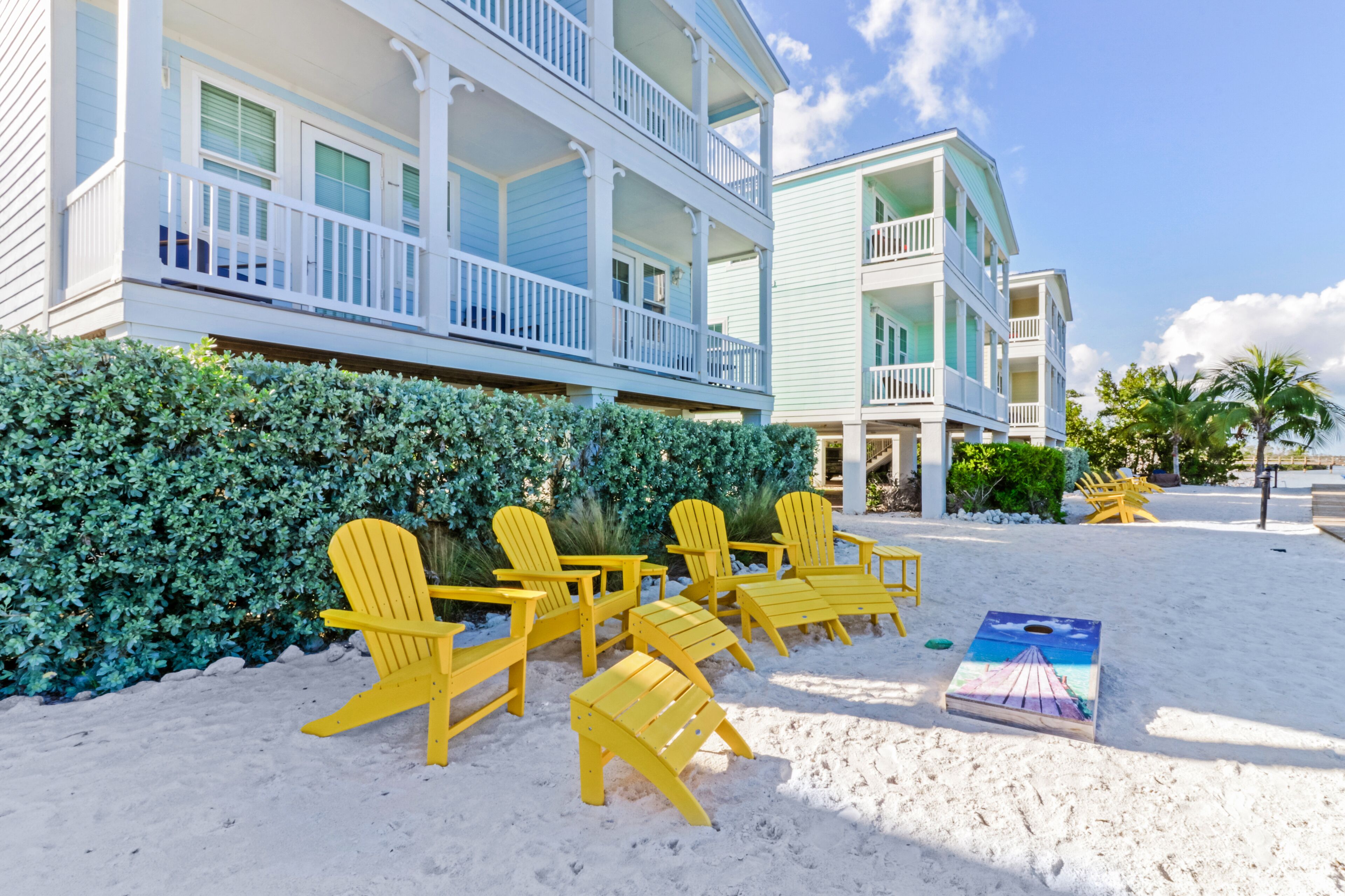 A beachfront hotel with multiple balconies, a sandy beach area with yellow chairs, and a hedge.