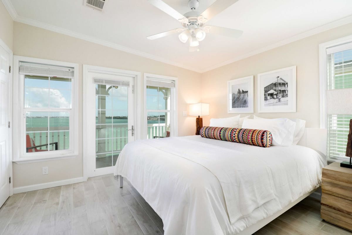 A bedroom with a bed, a ceiling fan, a window with a view, and a wall with framed pictures.