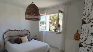 A bedroom with a bed, a hanging wicker light, a window with a view, and a bottle on the wall.