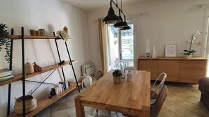 A dining area with a wooden table, white chairs, a black pendant light, and shelves with various items.