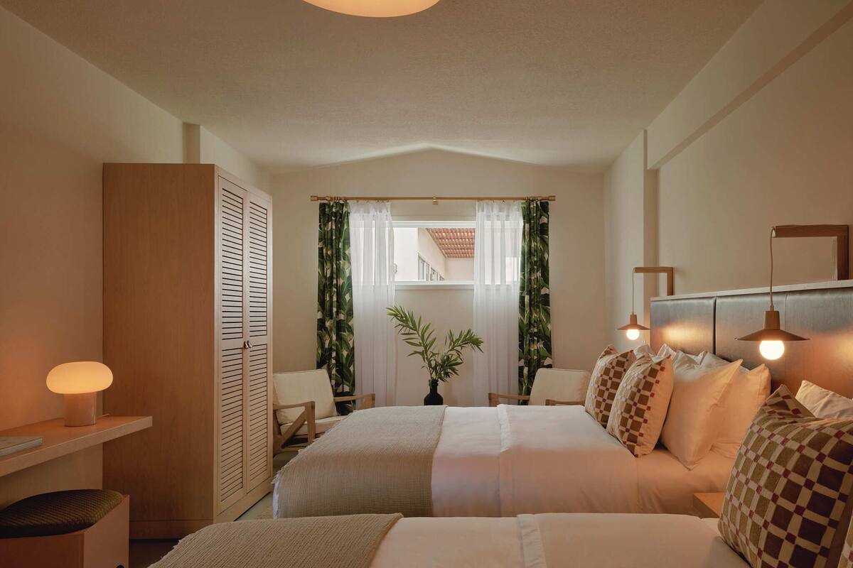 A hotel room with two beds, a wooden wardrobe, a desk with a lamp, and a view of a balcony.