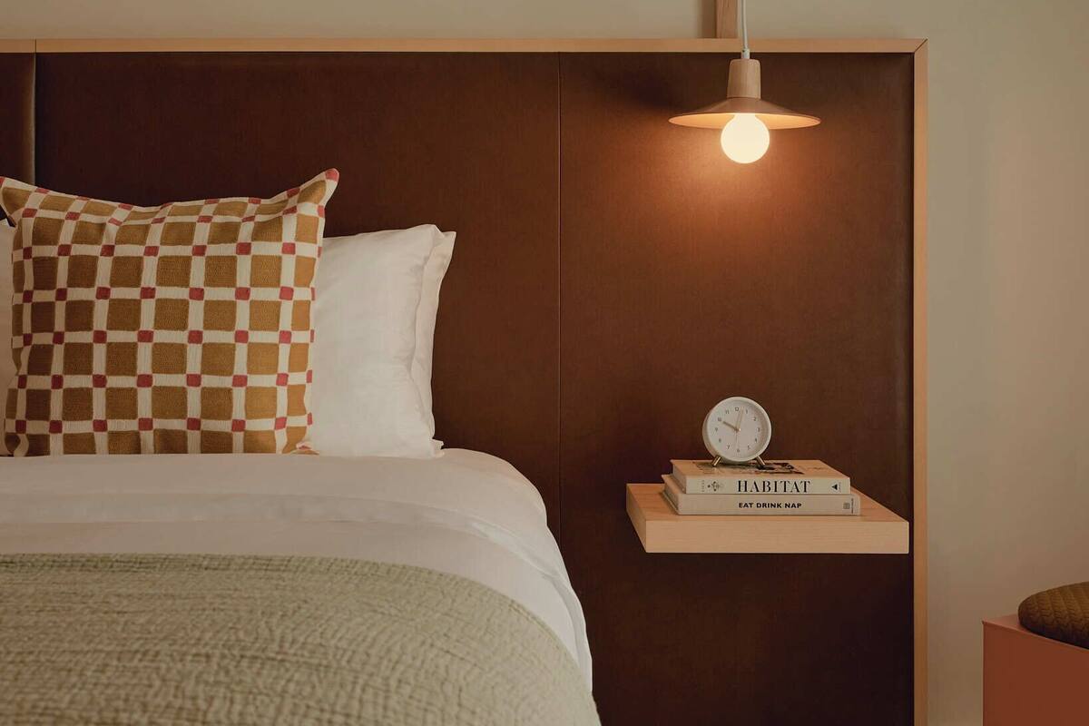 A neatly made bed with a checkered pillow, a bedside table with a clock and books, and a hanging light fixture.