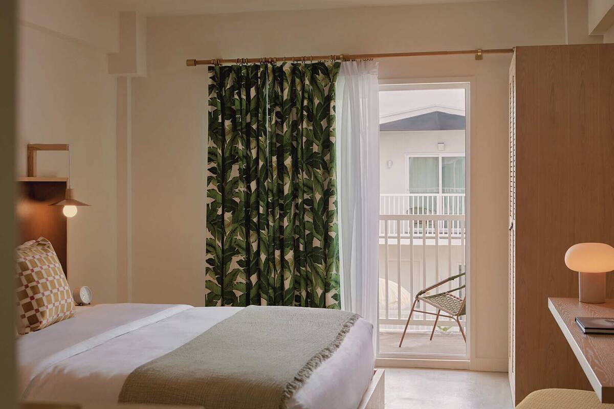 A hotel room with a bed, a desk, a chair, a balcony with a view, and a curtain with a leaf pattern.