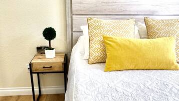 Ein ordentlich gemachtes Bett mit gemusterten Kissen, ein Nachttisch mit kleiner Topfpflanze und ein gerahmter Spruch an der Wand.