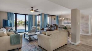 Ein geräumiges Wohnzimmer mit einer Couch, Sesseln und einem Couchtisch. Es gibt einen Deckenventilator und große Fenster mit Vorhängen.