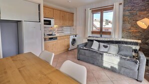A compact kitchen with wooden cabinets, a microwave, and a washing machine. There's a grey sofa with a heart design, a wooden dining table, and white chairs. A stone wall and a window with curtains are also visible. - 250m from ski slopes, 2nd floor, ski locker, 38m², Méribel-Mottaret (Méribel - Les Allues)