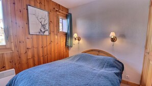 A bedroom with a wooden paneled wall, a bed with a blue quilt, two wall-mounted lamps, and a framed picture of a deer. - 250m from ski slopes, 2nd floor, ski locker, 38m², Méribel-Mottaret (Méribel - Les Allues)