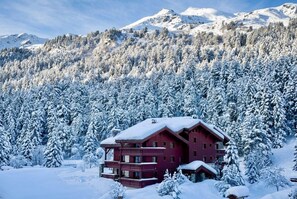 A multi-story chalet with a red facade, surrounded by snow-covered trees and mountains in the background.