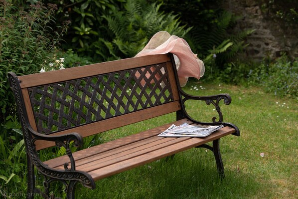 Un banc en bois doté d’un dossier ajouré et de supports d’accoudoirs en métal, placé sur une pelouse, avec un feuillage dense en arrière-plan.
