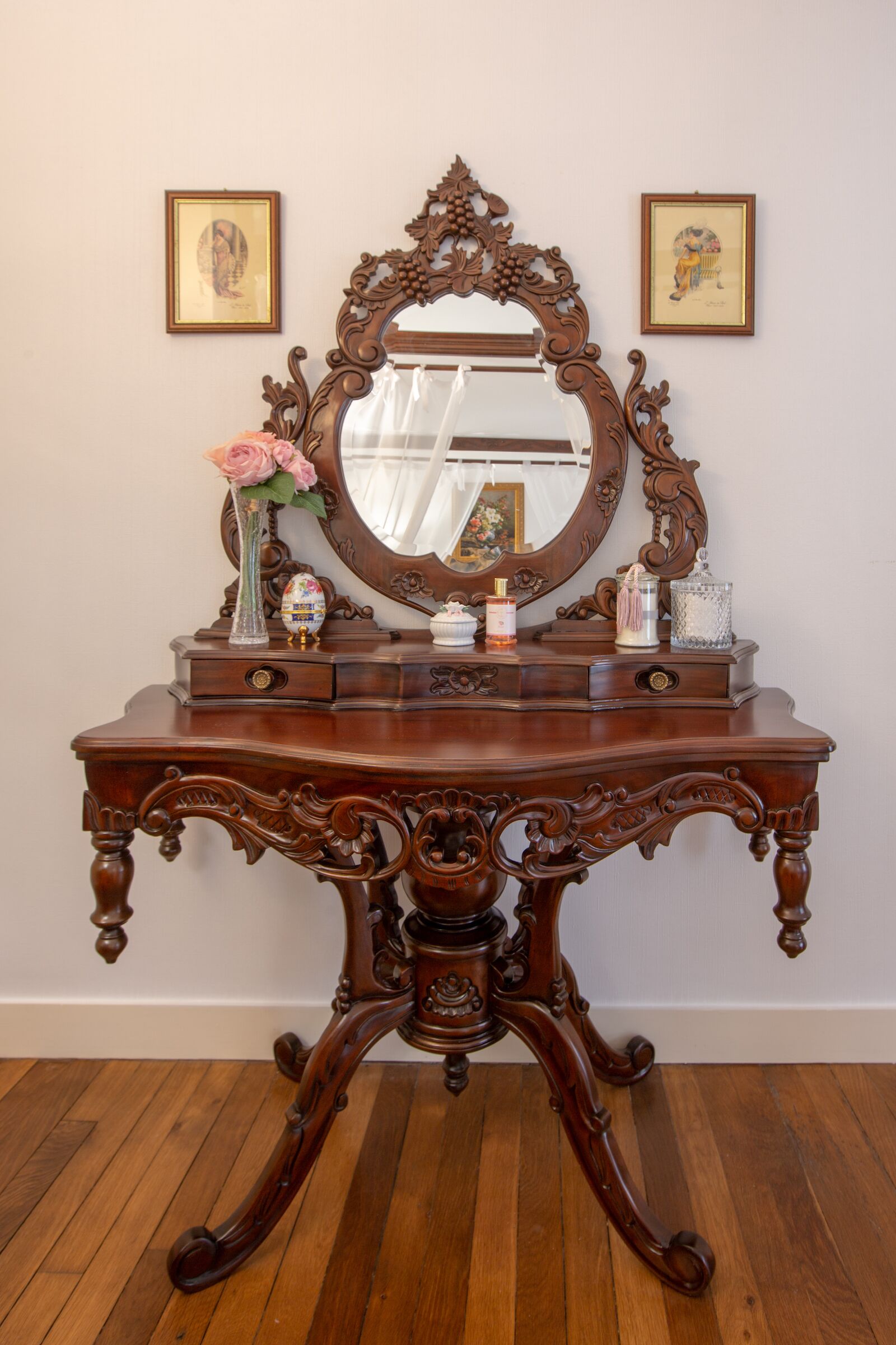 Une coiffeuse en bois avec un grand miroir, deux tableaux encadrés accrochés au mur et un vase rempli de fleurs posé sur la coiffeuse.
