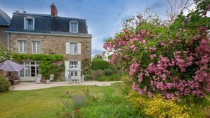 Une maison en pierre avec un jardin, une terrasse dotée de mobilier d’extérieur et un grand arbuste en fleurs.