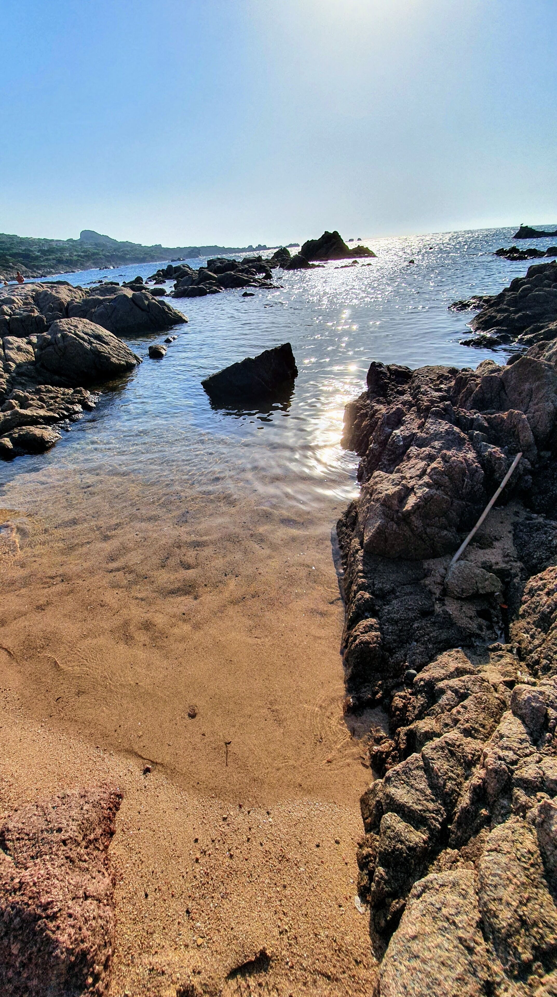 A rocky shoreline with clear water and a visible stick in the sand.