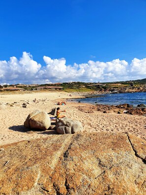 A beach with large rocks, a clear sky, and distant buildings.